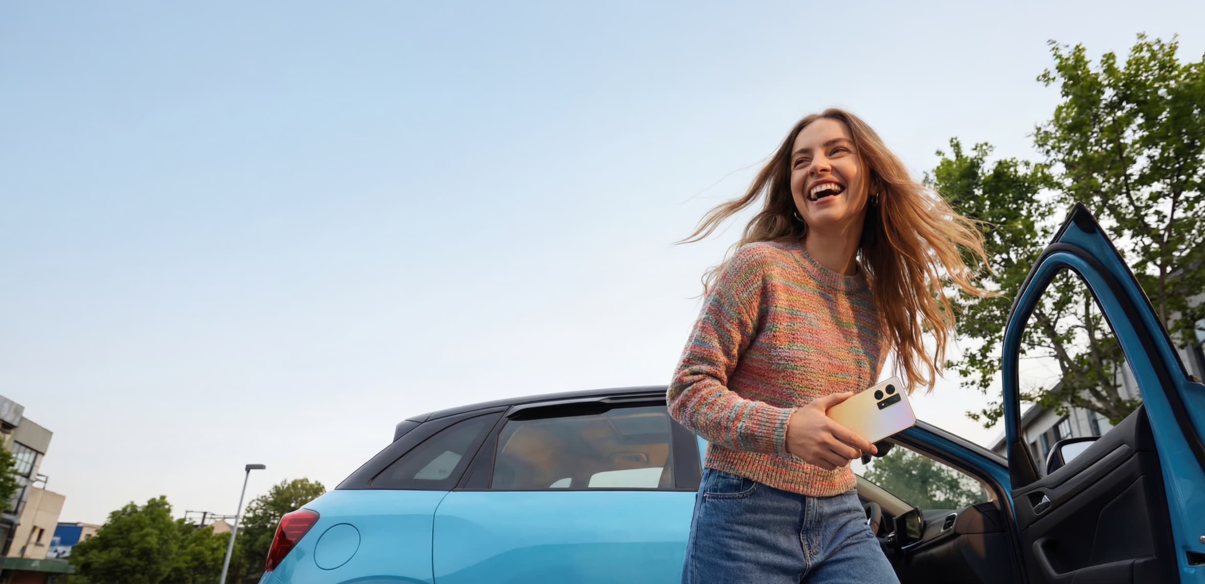 Eine lachende Person mit Handy in der Hand steigt aus einem blauen Auto