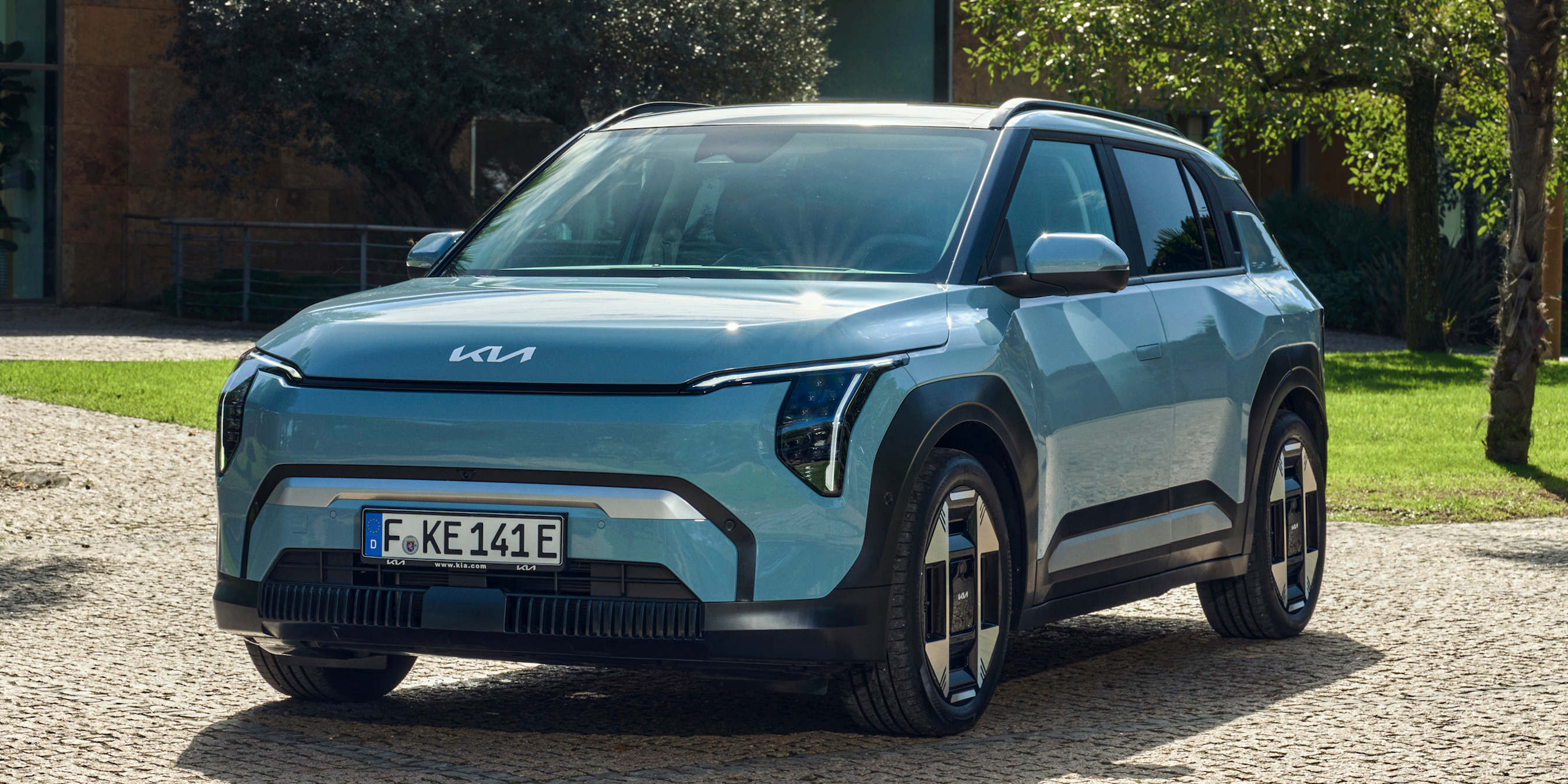 Das Bild zeigt einen hellblauen Kia EV SUV, der auf einem gepflasterten Hof vor einem Gebäude mit üppigem Grün im Hintergrund geparkt ist. Das Fahrzeug ist frontal leicht angeschnitten und der Fokus liegt auf dem Design des Autos.