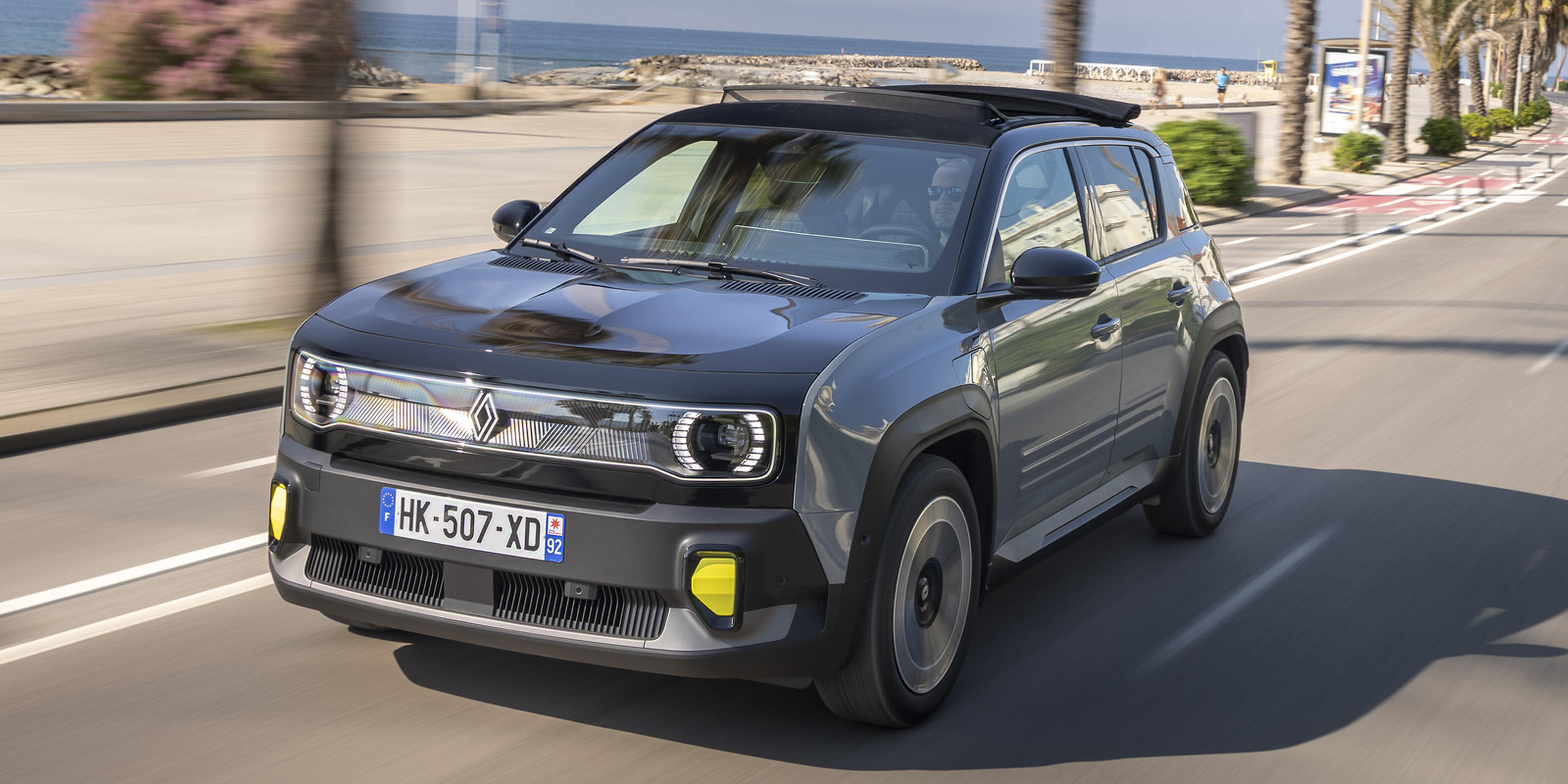 Ein schiefergrauer Renault 4 E-Tech elektrisch mit offenem Faltdach fährt an einer Strandpromenade entlang. Er hat ein französisches Kennzeichen.