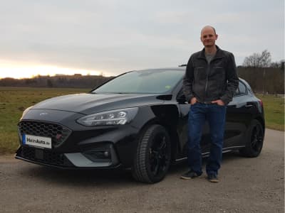 Ein Mann steht neben einem schwarzen Ford Focus ST auf einem Ländlichen Weg. Das Fahrzeug trägt das Logo von MeinAuto.de auf dem Kennzeichen.