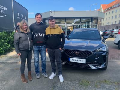 Eine Familie steht lächelnd vor ihrem dunklen Cupra Formentor SUV auf einem Autohaus-Gelände. Das Fahrzeug trägt ein MeinAuto.de-Kennzeichen.