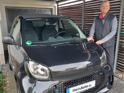 Ein Mann steht lächelnd neben einem schwarzen Smart EQ fortwo Cabrio unter einem Carport. Das Fahrzeug trägt ein MeinAuto.de-Nummernschild.