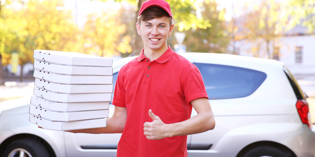 Junger Mann liefert Pizza mit einem Auto aus. Pizzakartons in der einen Hand, andere Hand Daumen hoch.