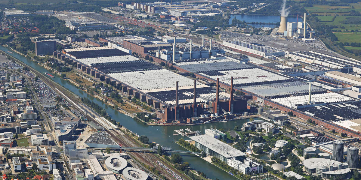 Das Bild zeigt eine Luftaufnahme des Volkswagen-Werks in Wolfsburg. Man sieht die riesige Ausdehnung des Werksgeländes mit seinen vielen Gebäuden und Produktionshallen, die sich entlang eines Kanals erstrecken.