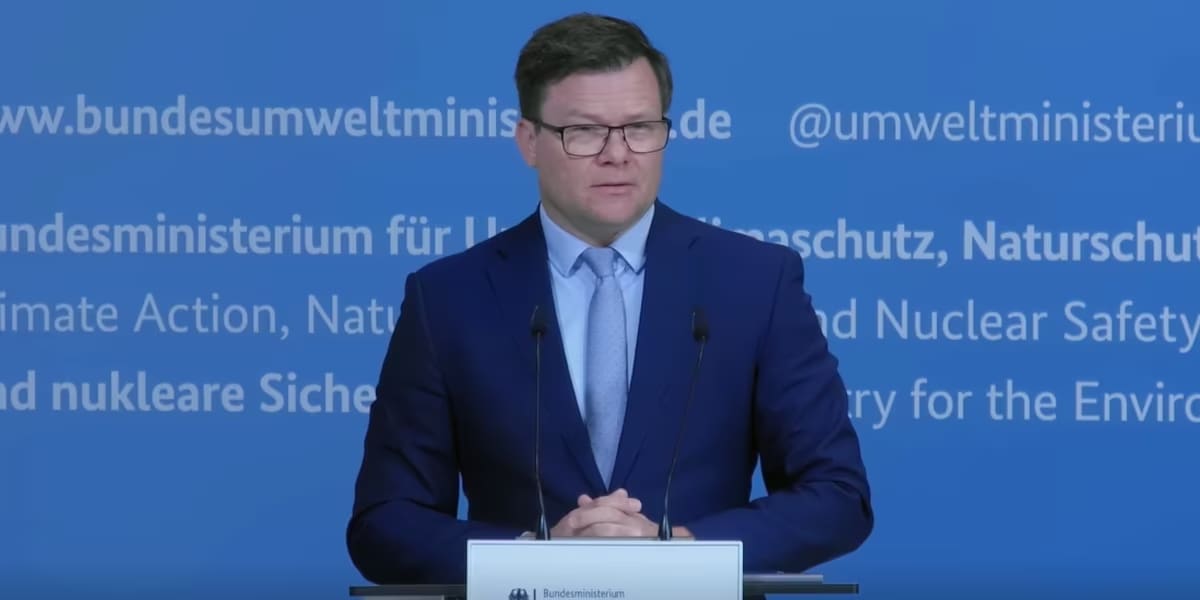 Ein Mann in einem dunkelblauen Anzug und hellblauer Krawatte steht hinter einem Rednerpult. Er trägt eine Brille und hat die Hände vor sich gefaltet. Im Hintergrund befindet sich eine blaue Wand.
