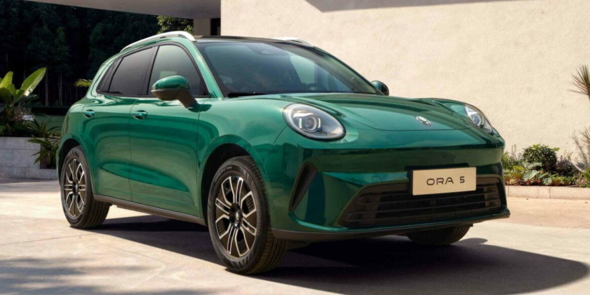 Ein kompakter, metallic-grüner GWM Ora 5 SUV mit runden Scheinwerfern steht auf einer sonnigen Einfahrt vor einem modernen Wohnhaus mit heller Fassade und Bepflanzung im Hintergrund.
