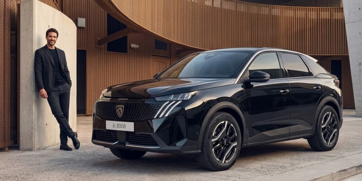 Ein lächelnder Mann im eleganten, dunklen Anzug lehnt an einer Betonwand neben einem glänzenden, schwarzen Peugeot E-3008 SUV vor einer modernen, braun strukturierten Gebäudefassade.
