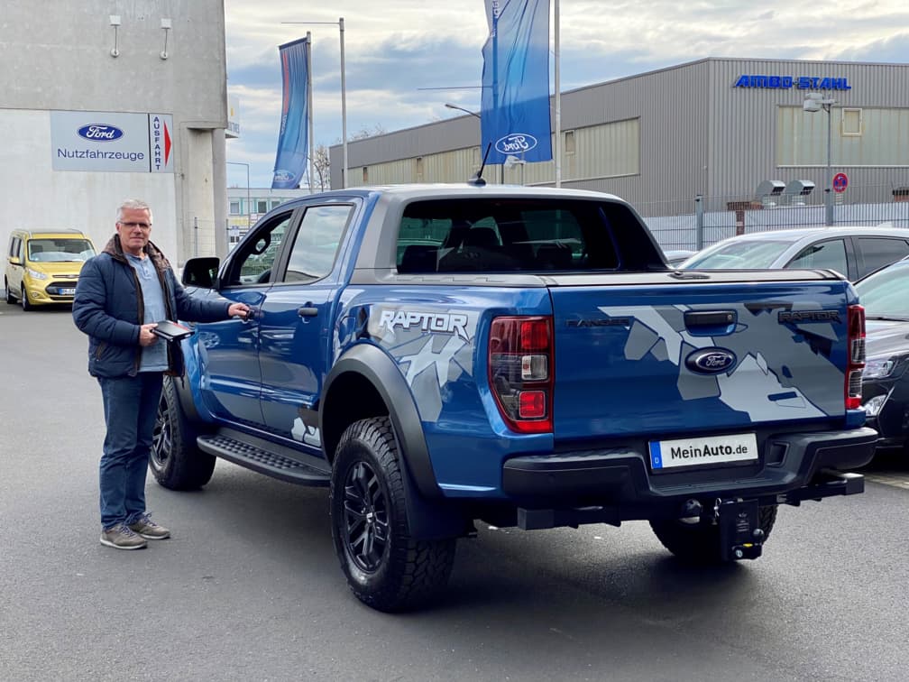 Ein Mann steht vor einem blauen Ford Ranger Raptor Pickup auf dem Gelände eines Autohauses. Der Geländewagen trägt auffällige Grafiken auf der Ladefläche.