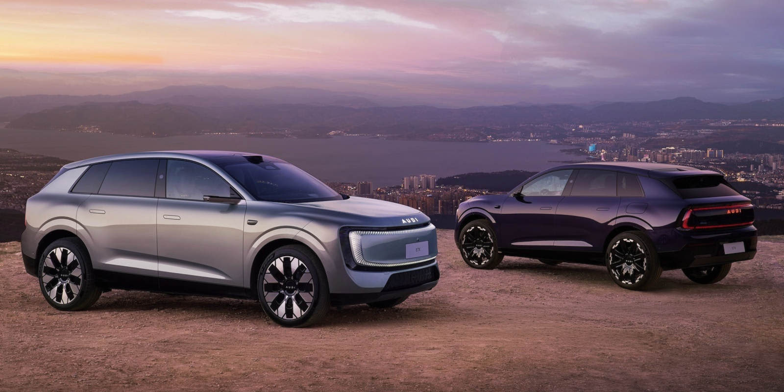 Zwei moderne Audi E7X Elektro-SUVs (silber & dunkelblau) stehen bei Sonnenuntergang auf einem Hügel über einer Küstenstadt. Das futuristische Design zeigt markante LED-Lichtsignaturen und großflächige, geschlossene Frontpartien.