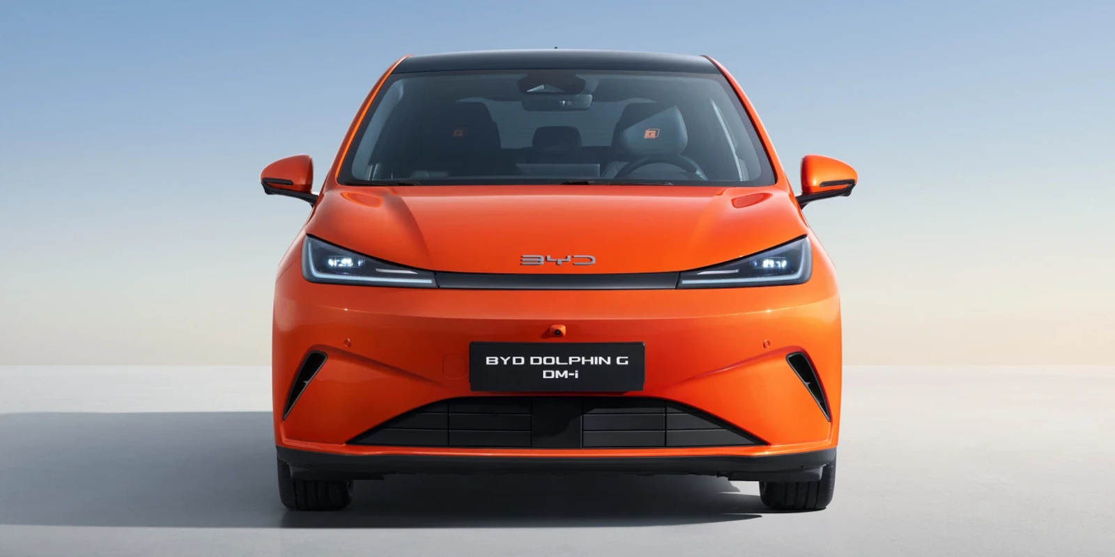 Ein orangefarbener BYD Dolphin G DM-i ist von vorne vor hellem Hintergrund abgebildet. Das Kompaktfahrzeug hat ein schwarzes Dach, schmale Scheinwerfer und trägt die Modellbezeichnung auf dem Schild.