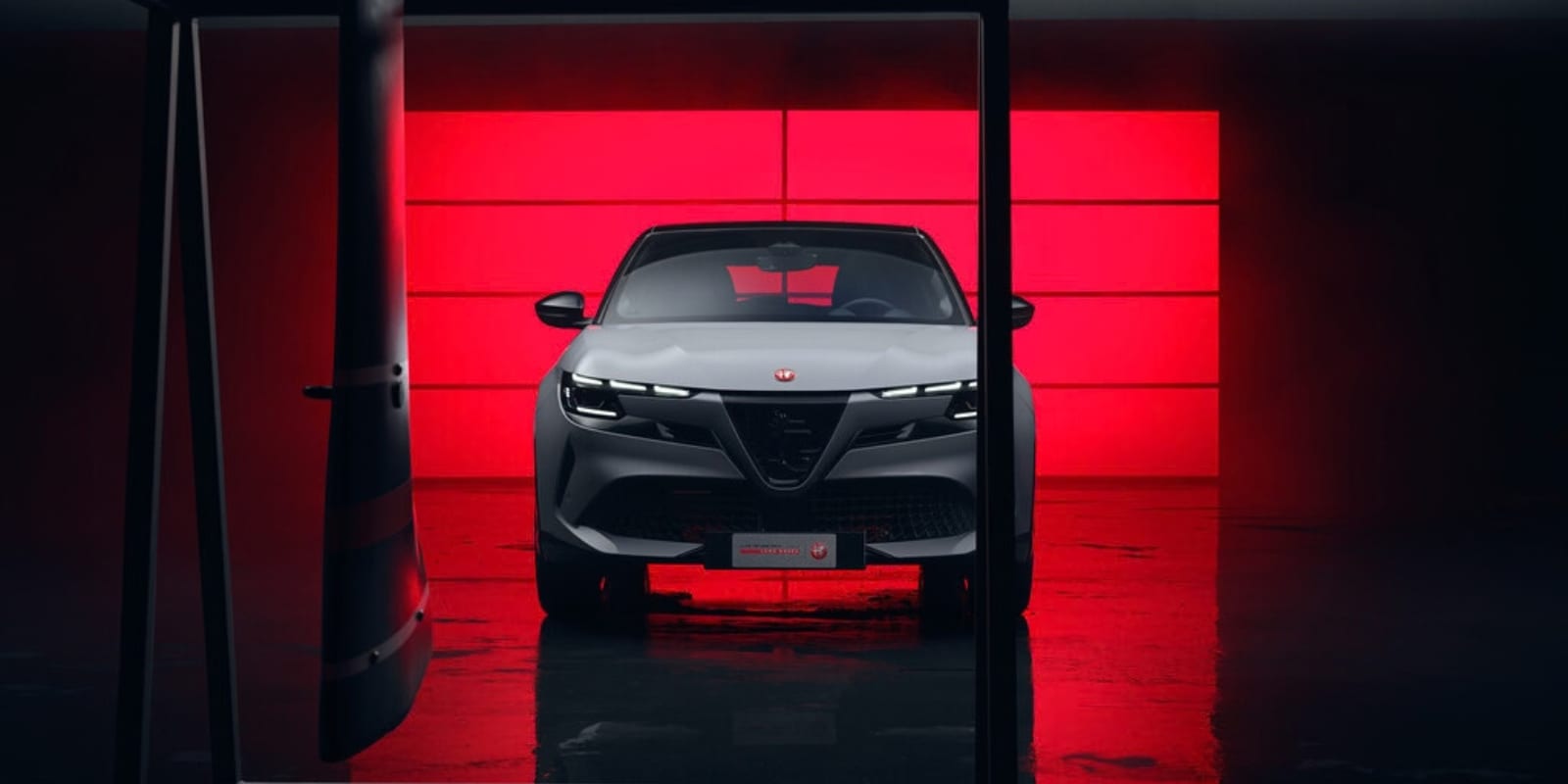 Ein grauer Alfa Romeo Junior steht vor einem rot leuchtenden Hintergrund. Prägend sind der dreieckige Scudetto-Grill und die dreiteiligen LED-Scheinwerfer.
