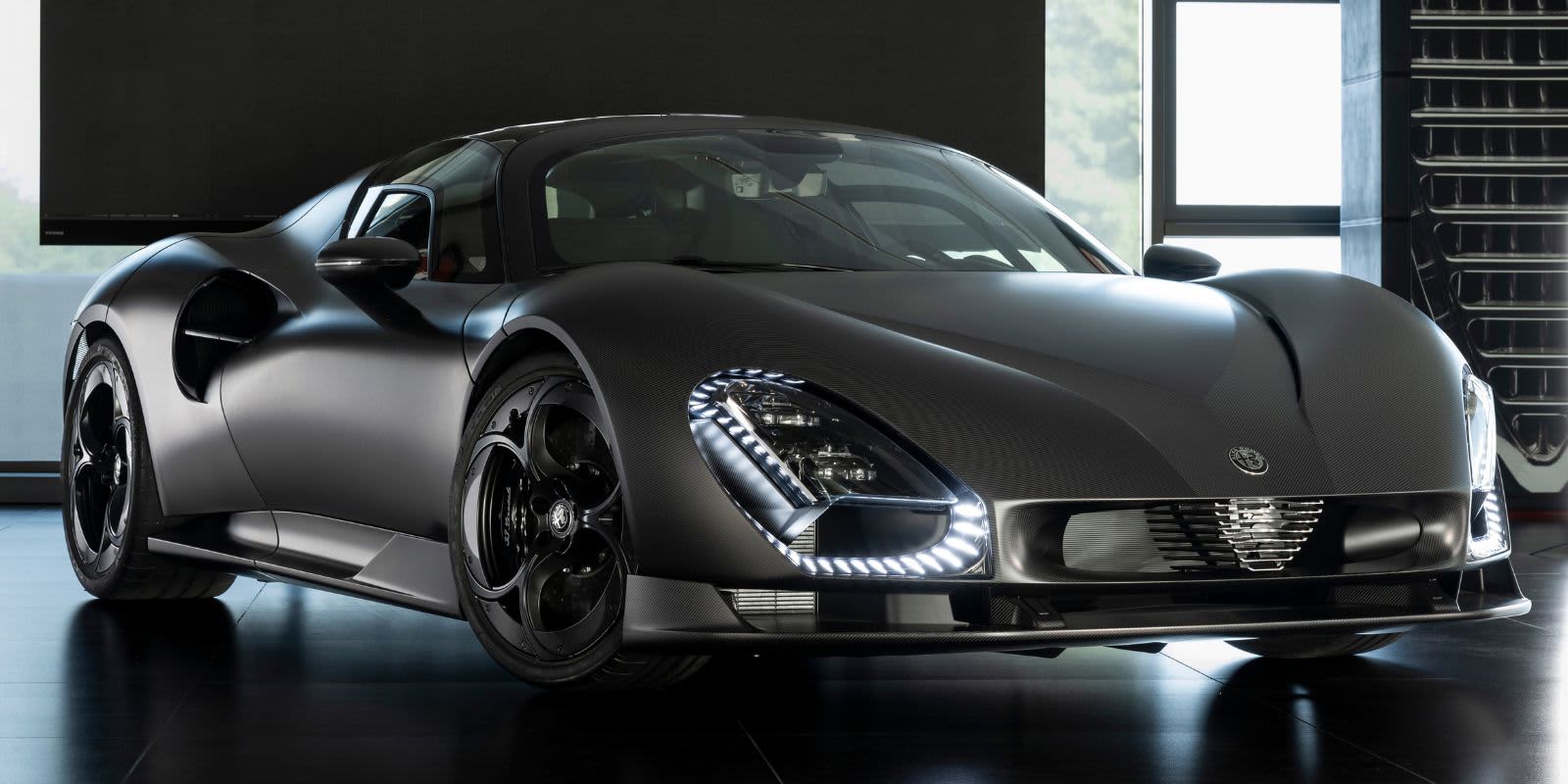 Ein mattgrauer Alfa Romeo 33 Stradale steht in einem modernen Studio. Prägend sind die ovalen LED-Scheinwerfer und die aerodynamisch geschwungene Karosserie.   