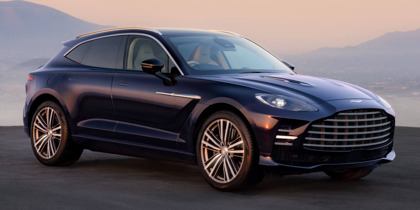 Ein dunkelblauer Aston Martin DBX steht bei Dämmerung vor Bergkulisse. Prägend sind der große Chrom-Kühlergrill und die elegante SUV-Silhouette.   
