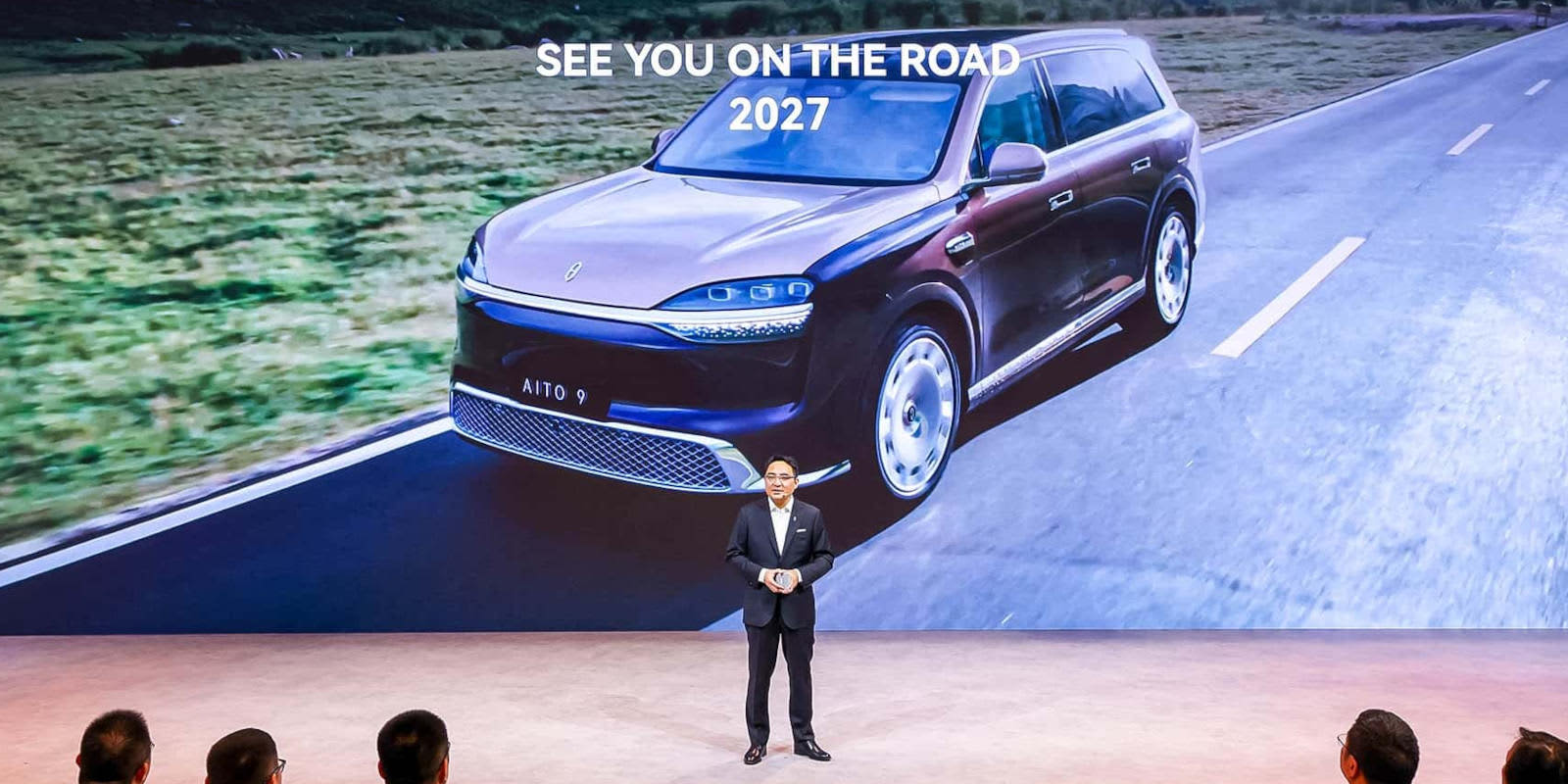 Ein Mann präsentiert den Aito 9 bei einer Messe vor einer großen Leinwand. Auf der Anzeige ist das Luxus-SUV auf einer Straße mit LED-Lichtern zu sehen.   