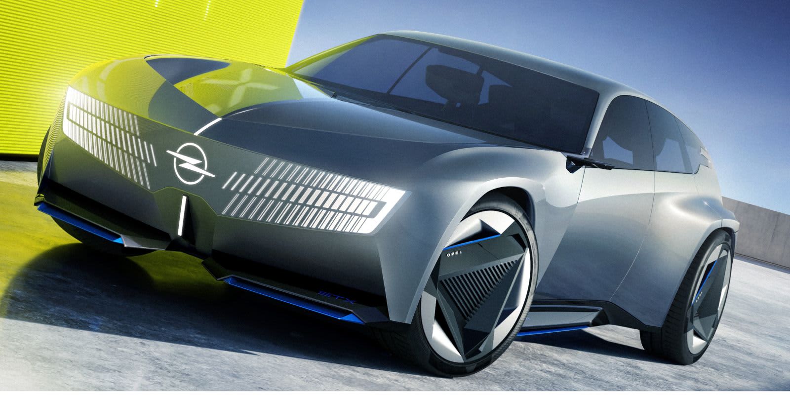 Ein graues Opel Concept Car steht draußen vor gelber Kulisse. Ins Auge fallen der leuchtende Vizor-Grill, scharfe Linien und auffällig gestaltete Felgen.