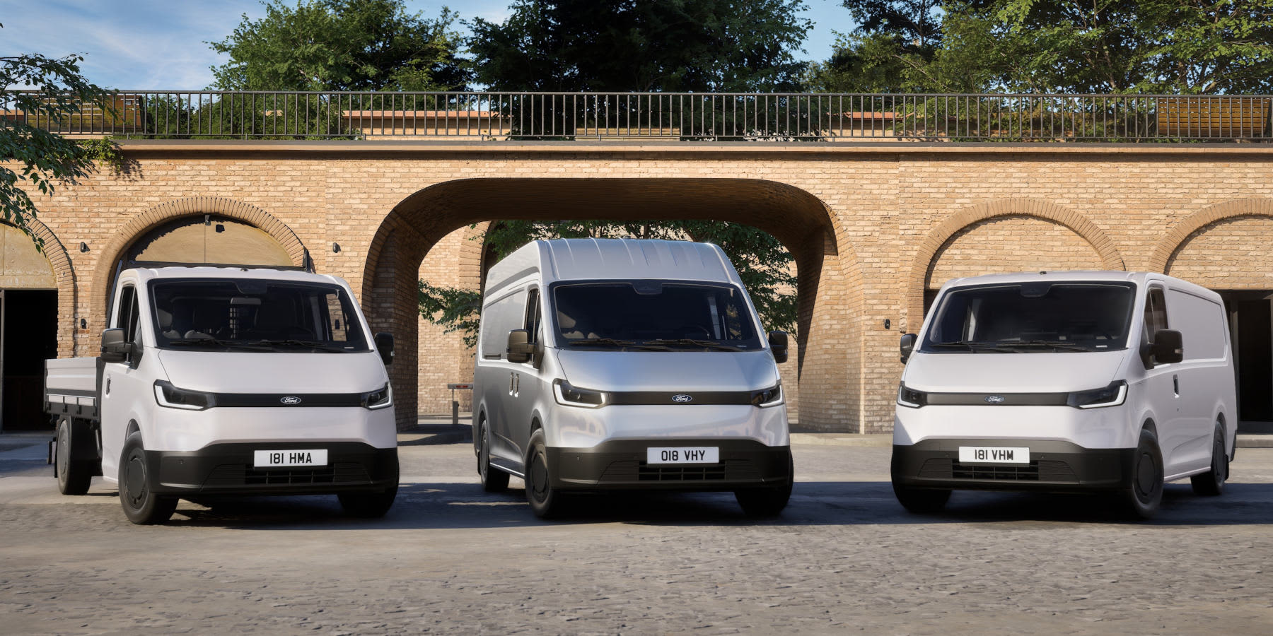 Drei weiße und silberne Ford Transit Nutzfahrzeuge – ein Pritschenwagen, ein Hochdach-Kastenwagen und ein klassischer Kastenwagen – stehen nebeneinander vor einer historischen Ziegelstein-Bogenarchitektur.