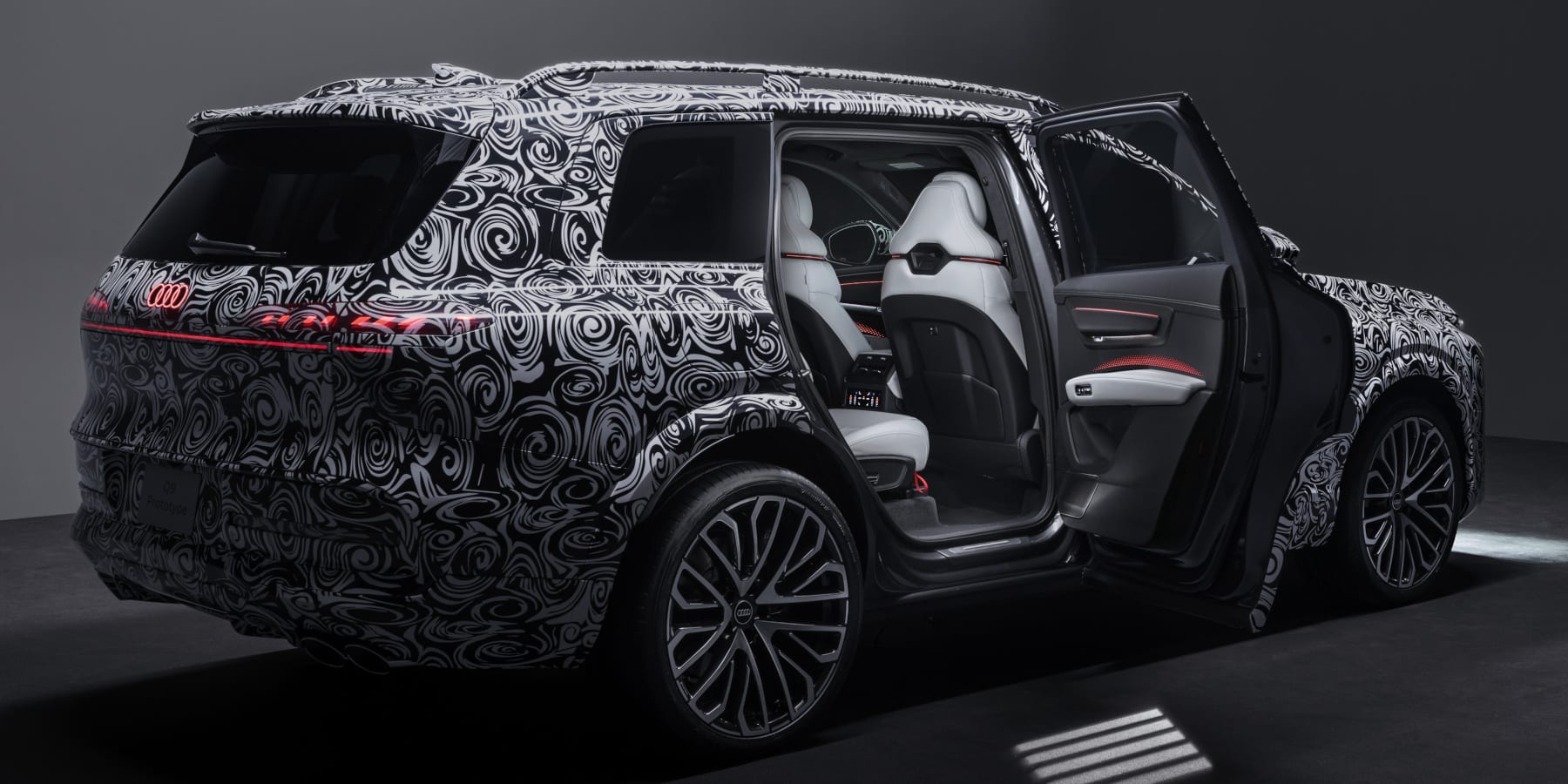 Ein getarnter Audi Q9 Prototyp mit markanter schwarz-weißer Folierung. Das große SUV zeigt geöffnete Türen, ein beleuchtetes rotes Logo am Heck und einen luxuriösen hellen Innenraum.