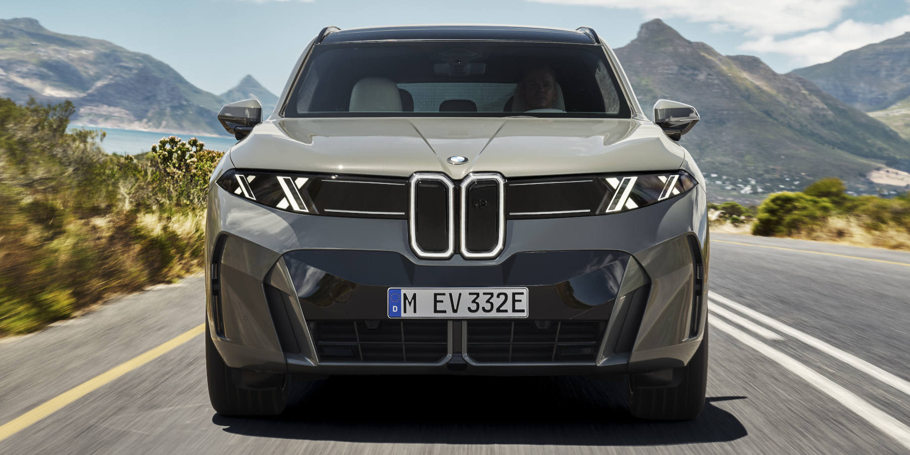 Frontansicht eines grauen BMW-Elektro-SUVs (Neue Klasse), das auf einer Küstenstraße fährt. Das Fahrzeug markiert sich durch eine beleuchtete, schmale Doppelniere, Scheinwerfer mit doppelten X-förmigen Leuchtelementen.