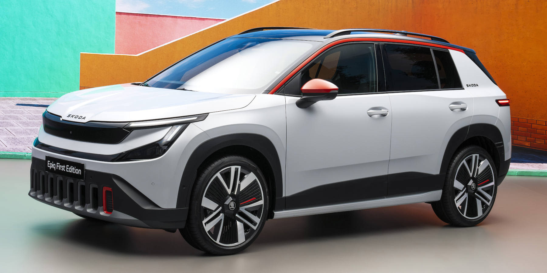 Ein kompakter, weißer Skoda Epiq SUV mit schwarzem Dach und orangefarbenen Akzenten an Spiegeln und Frontschürze steht schräg vor einem farbenfrohen, modern gestalteten Hintergrund.