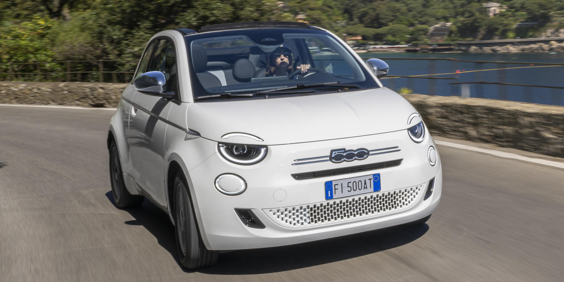 Ein weißer Fiat 500 Dolcevita fährt mit geöffnetem Verdeck auf einer Küstenstraße. Er hat verchromte Außenspiegel und trägt das italienische Kennzeichen FI 500AT.