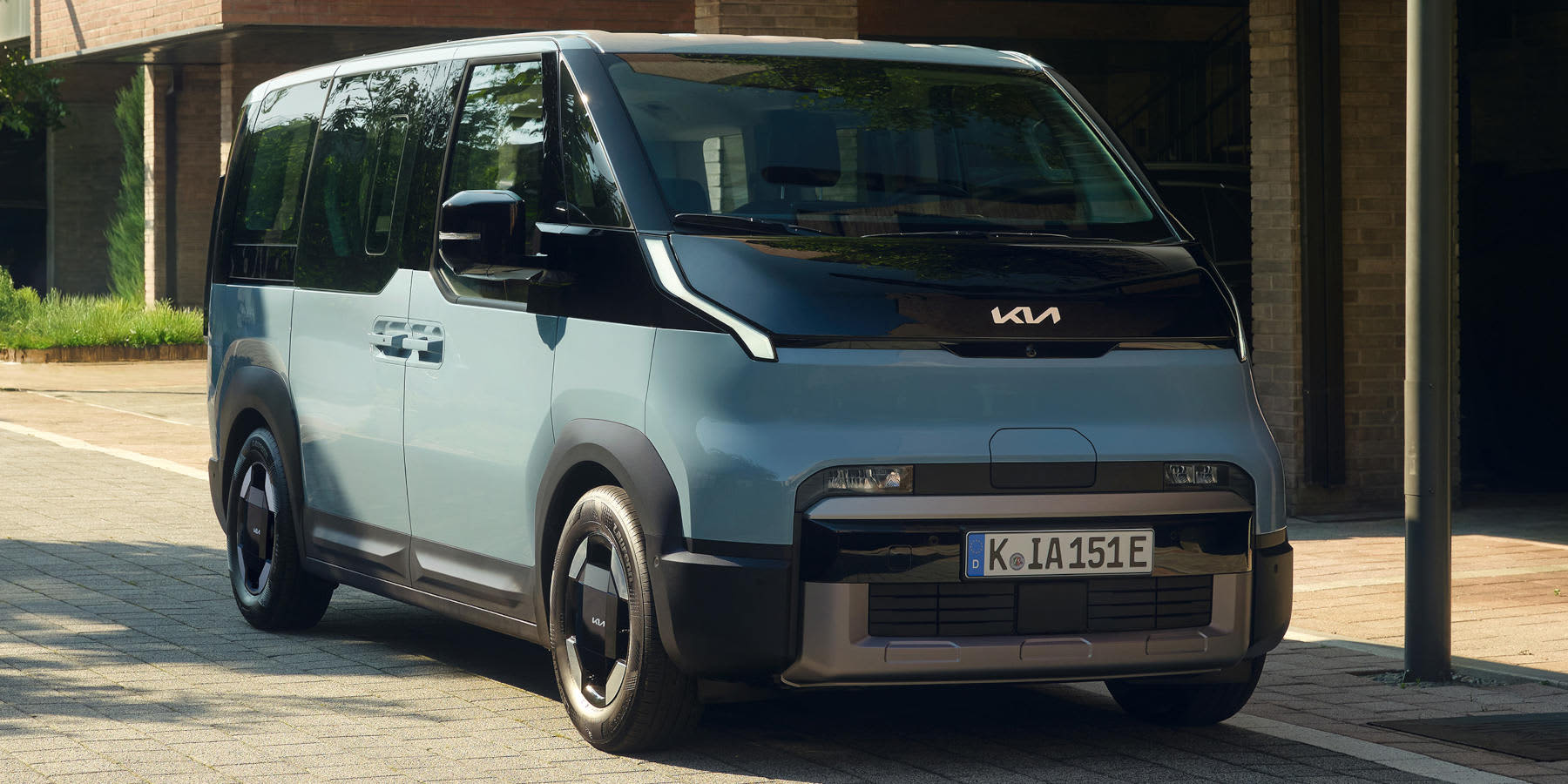 Ein hellblauer, futuristischer Kia PV5 Passenger Elektro-Van steht auf einem gepflasterten Weg. Er hat eine markante schwarze Frontpartie, aerodynamische Felgen und das deutsche Kennzeichen K IA 151E.