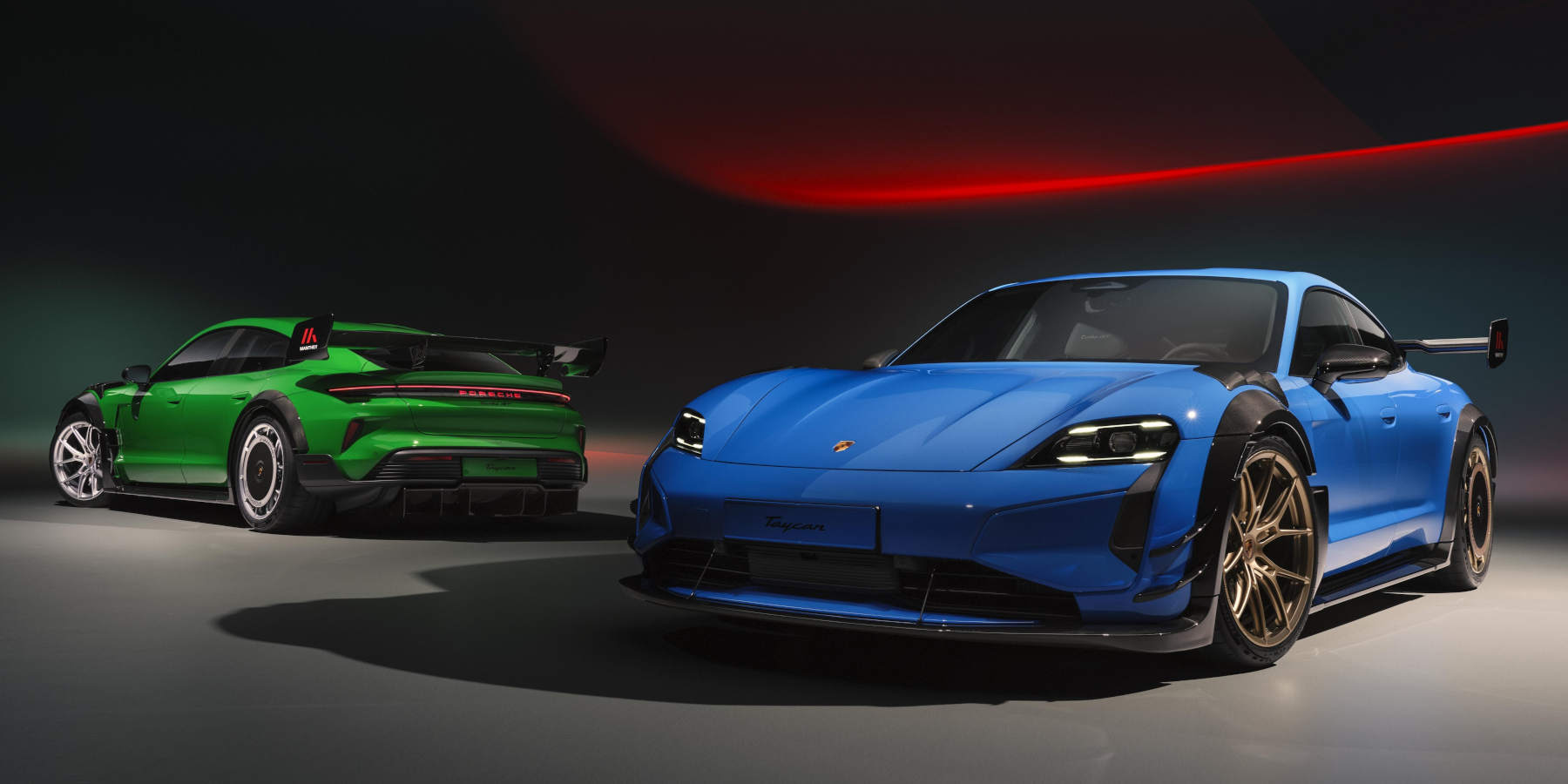 Zwei Porsche Taycan Turbo GT mit extremen Manthey-Rennsport-Kits (ein blauer vorne, ein grüner im Hintergrund) präsentieren sich mit großen Heckflügeln vor dunklem Hintergrund.