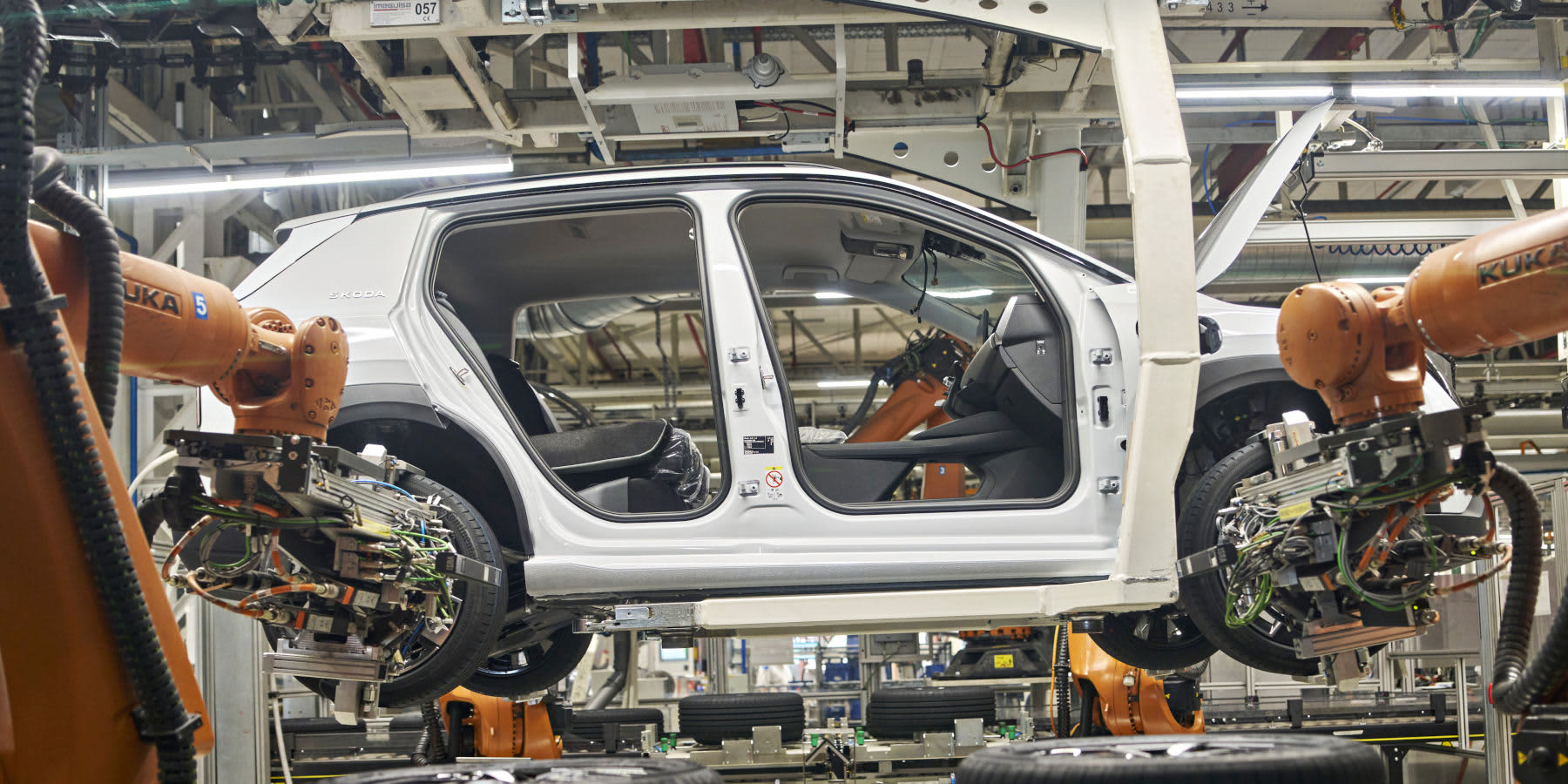 Die Rohkarosse eines weißen Škoda Epiq befindet sich in einer Fabrik auf dem Fließband. Orangefarbene Kuka-Montageroboter arbeiten an der Fertigung des kompakten Elektro-SUVs.