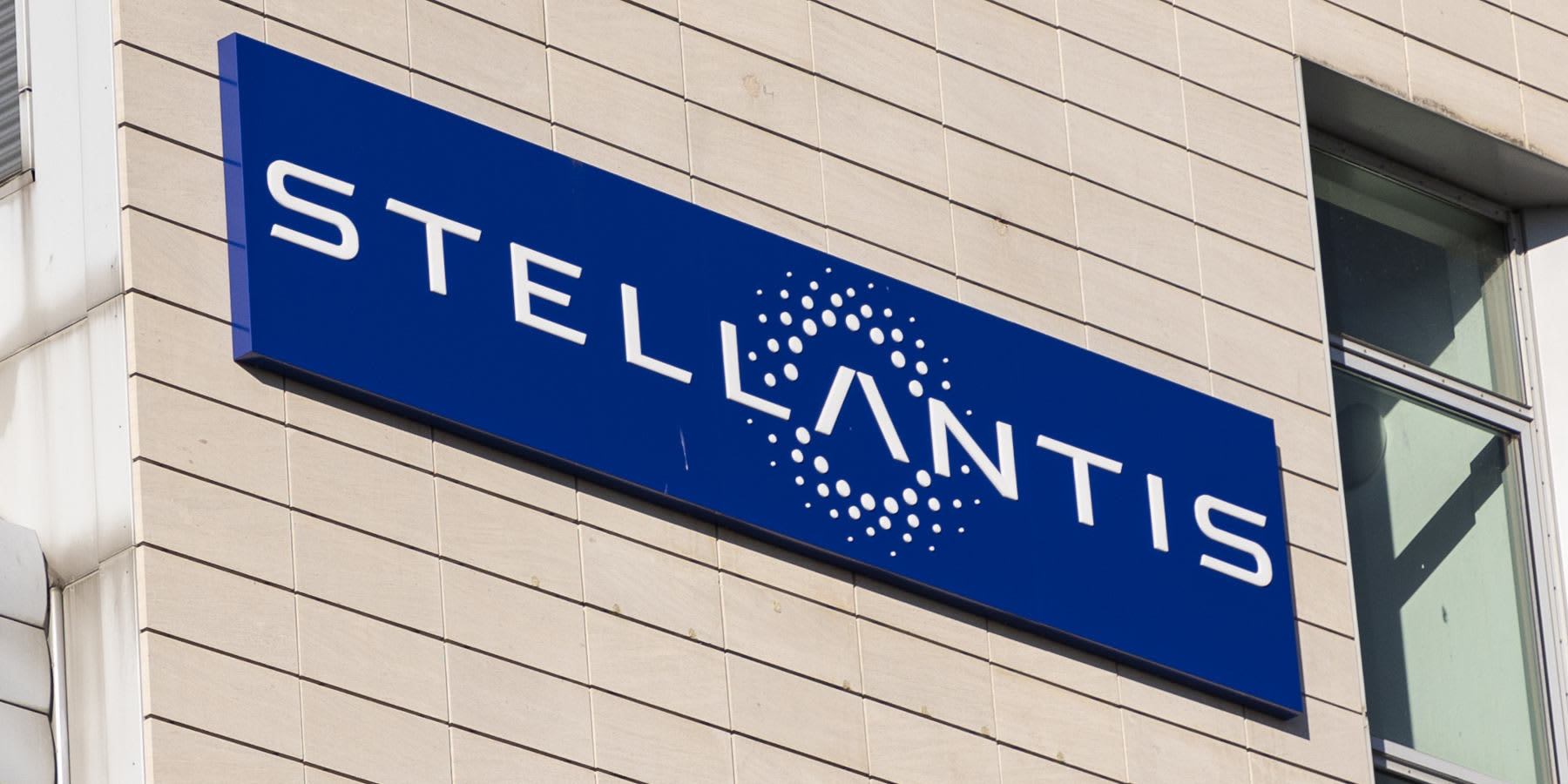 Ein blaues, rechteckiges Firmenschild mit dem weißen Schriftzug und Logo von „STELLANTIS“ ist schräg an einer hellen, gefliesten Fassade eines Gebäudes angebracht.