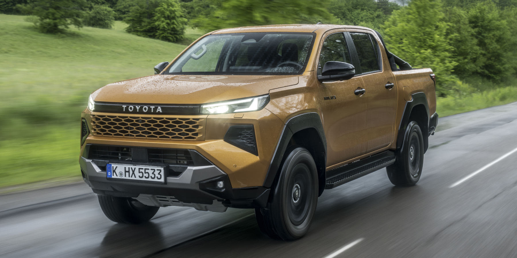 Ein gold-oranger Toyota Hilux Pick-up fährt auf einer nassen Landstraße an einer grünen Wiese vorbei. Das Fahrzeug hat eine Doppelkabine, schwarze Akzente und ein Kölner Kennzeichen (K HX 5533).