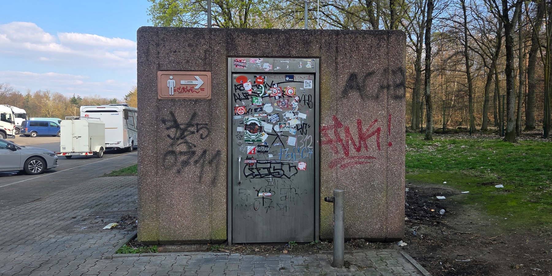 Ein heruntergekommenes Toilettenhäuschen aus Beton an einem Rastplatz. Die Metalltür und die Wände sind stark mit Graffitis und vielen Stickern beschmiert.