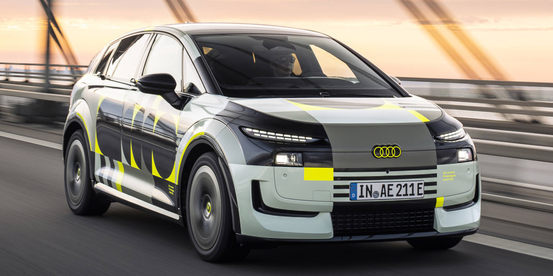 Ein getarnter Audi A2 e-tron Erlkönig fährt auf einer Brücke bei Sonnenuntergang. Auffällig sind neongelbe Akzente und das Logo an der Front.