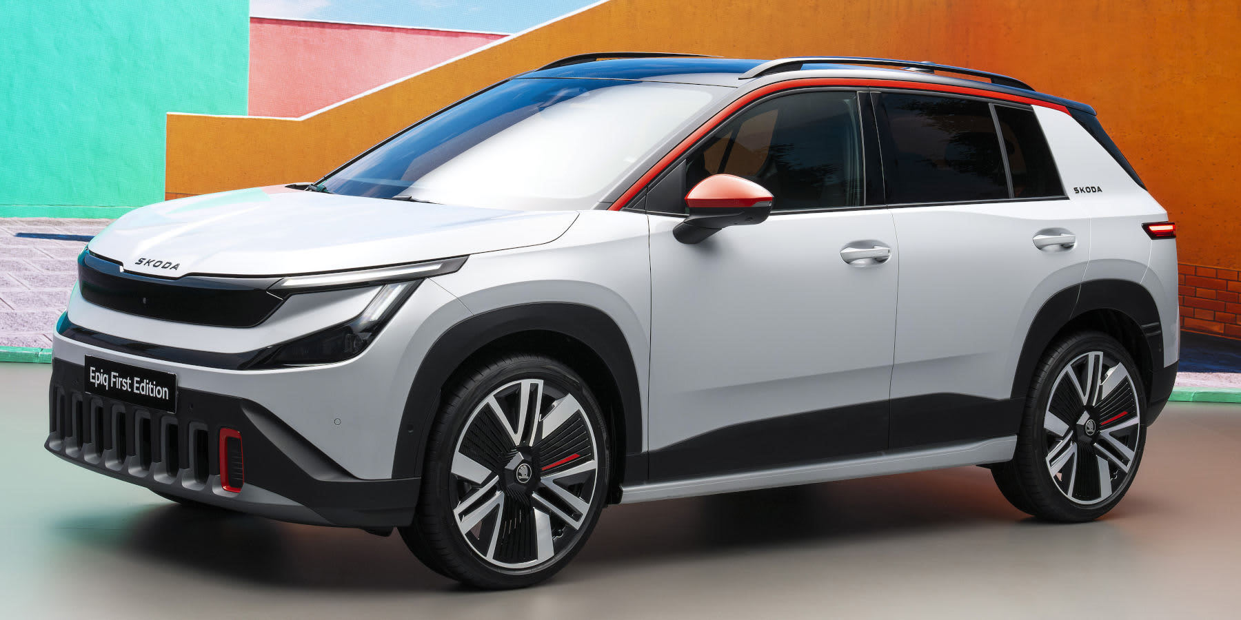 Ein weißer Škoda Epiq (First Edition) steht vor buntem Hintergrund. Das Kompakt-SUV hat orangefarbene Akzente an Dachlinie, Spiegeln und Grill.