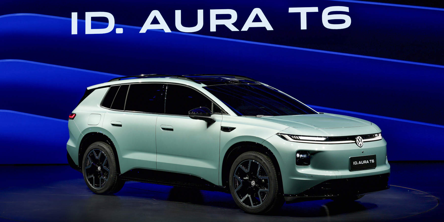 Ein mintgrüner VW SUV steht im Rampenlicht einer Bühne. Im Hintergrund prangt auf blauem Grund der Schriftzug „ID. AURA T6“, der auch am Kennzeichen steht.