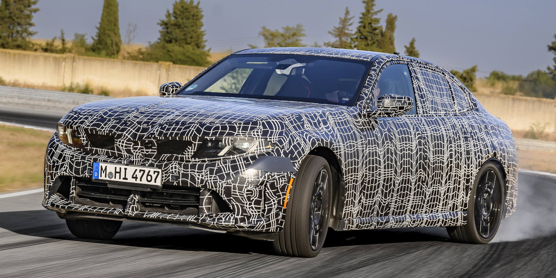 Ein stark tarnfolierter BMW 3er Erlkönig driftet dynamisch über eine Teststrecke, während Rauch von den Reifen aufsteigt.