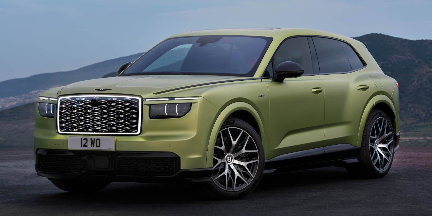 Ein olivenfarbener Bentley steht vor einer Hügellandschaft. Prägend sind der imposante, beleuchtete Kühlergrill und die schmalen Doppel-LED-Scheinwerfer.