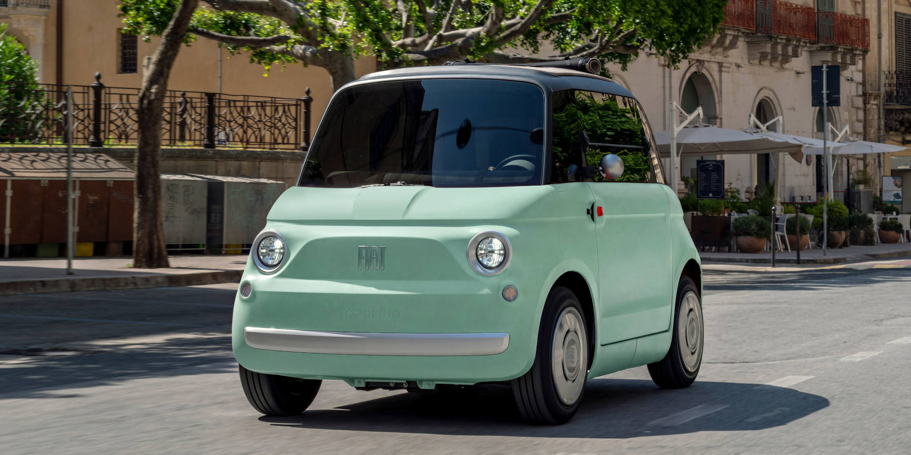 Ein mintgrüner Fiat Topolino Elektro-Kleinstwagen fährt durch eine sonnige Stadtstraße. Prägend sind das Retro-Design und die Rundleuchten.