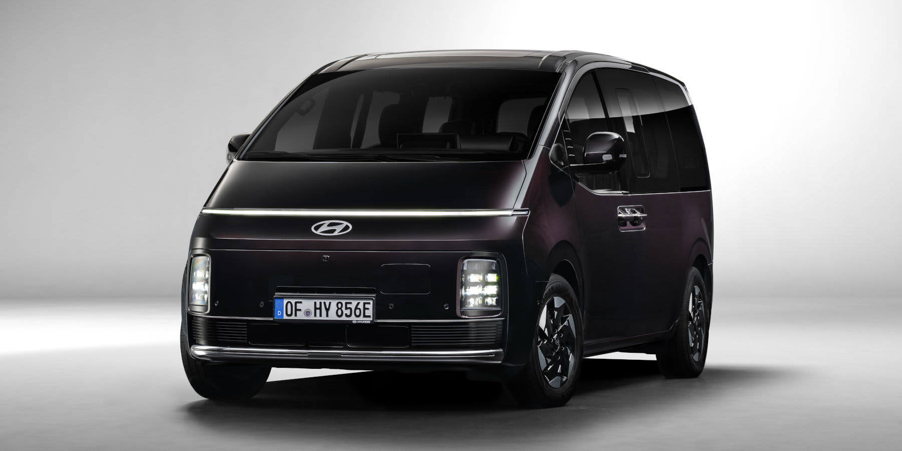 Ein dunkler Hyundai Staria Electric Großraumvan steht vor hellem Grund. Auffällig sind das durchgehende LED-Lichtband und die tief sitzenden Scheinwerfer.