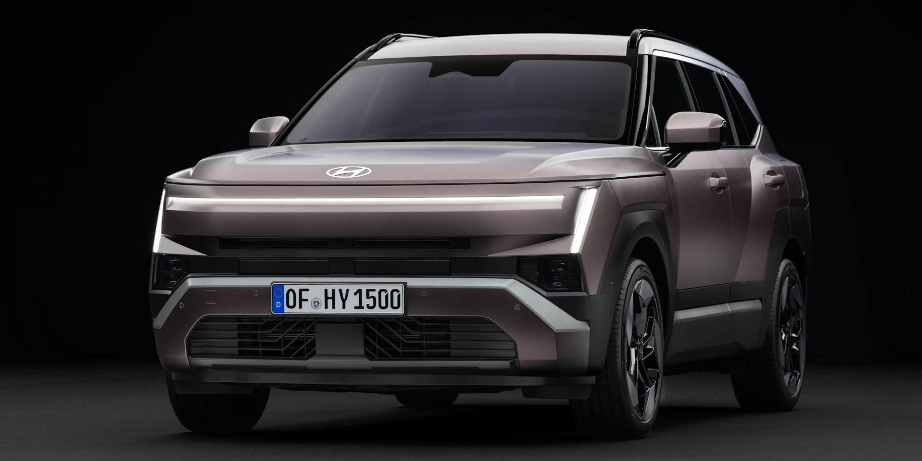 Ein metallicbrauner Hyundai steht im Studio auf dunklem Grund. Prägend sind das durchgehende LED-Lichtband an der Front und die markante, kantige SUV-Form.