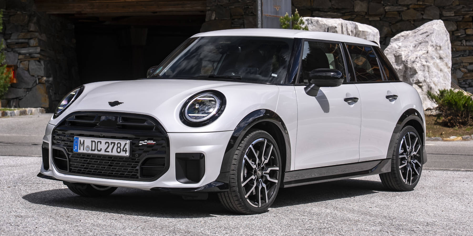 Ein weißer MINI Cooper S 5-Türer steht vor Fels- und Holzarchitektur. Markant sind die rundlichen LED-Scheinwerfer und das John Cooper Works Badge im Grill.