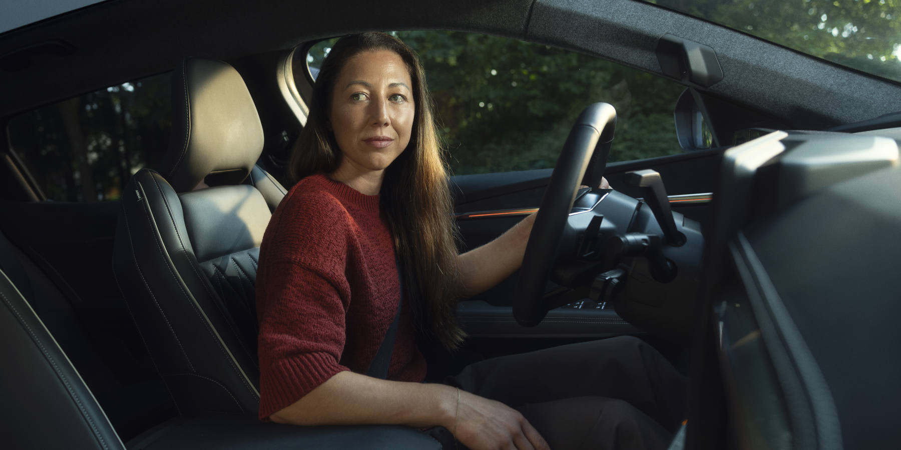 Eine Frau im roten Pullover sitzt am Steuer eines modernen Autos. Prägend sind das Lederinterieur, das Multifunktionslenkrad und das Cockpit-Design.