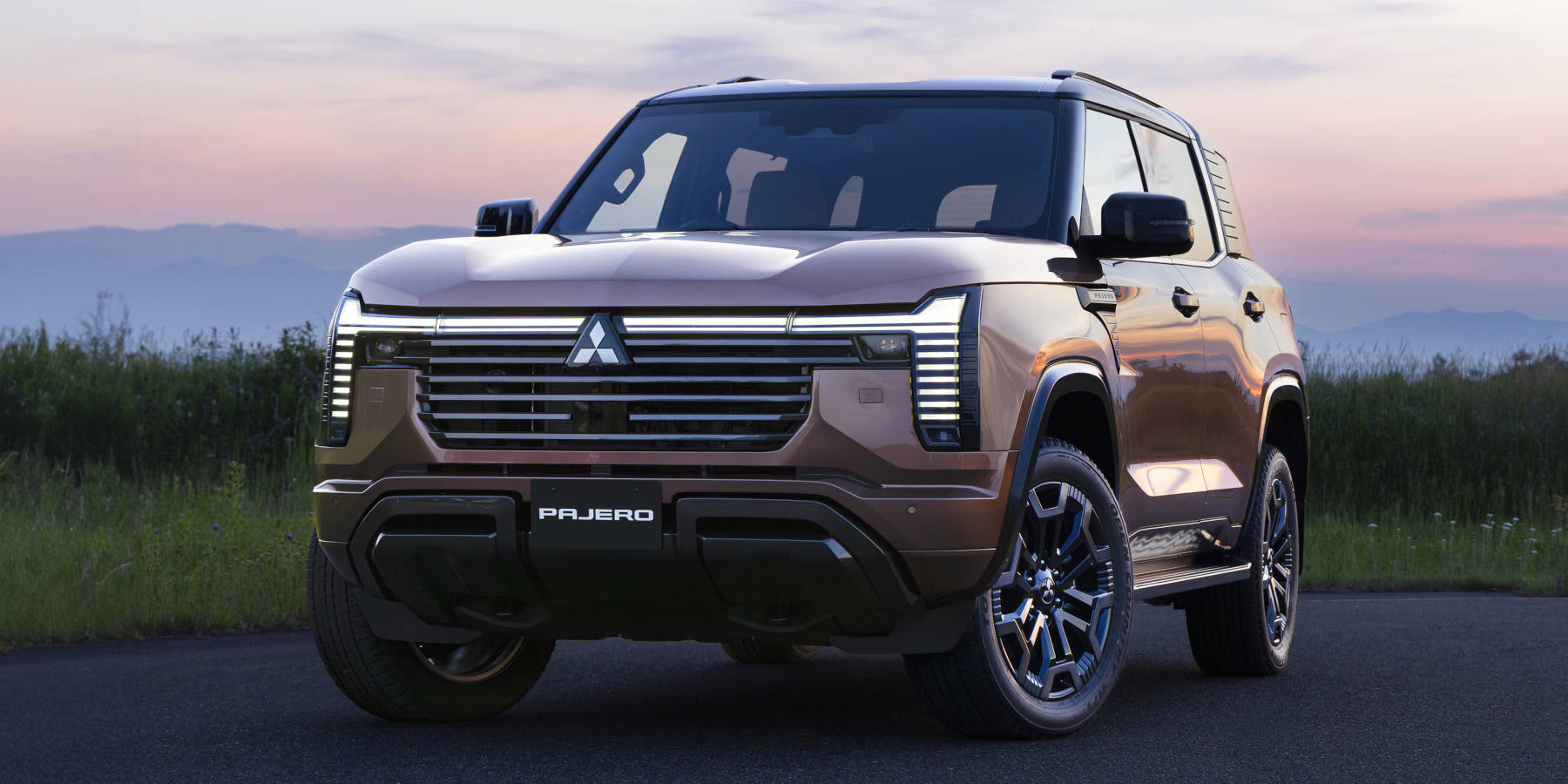 Ein brauner Mitsubishi Pajero Geländewagen steht auf einer Straße bei Dämmerung. Auffällig sind die breite Frontpartie mit LED-Lichtleiste und der Pajero-Schriftzug.