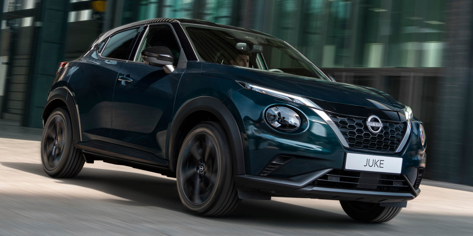 Ein dunkler Nissan Juke Pulse fährt durch eine urbane Kulisse. Prägend sind der V-Motion-Kühlergrill, runde LED-Scheinwerfer und die kompakte Crossover-Form.   