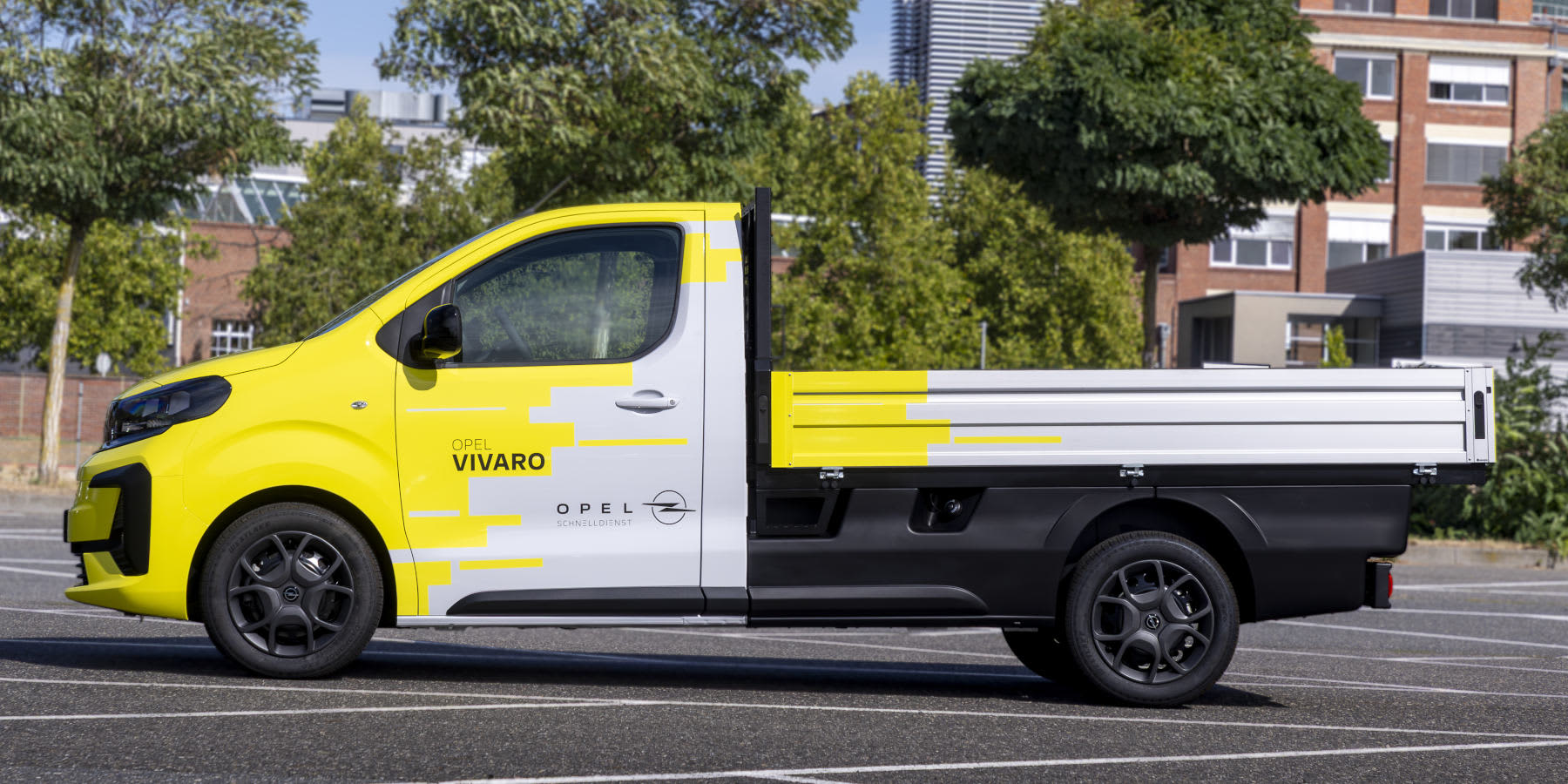 Ein gelb-weiß-schwarzer Opel Vivaro Pritschenwagen steht in der Seitenansicht auf einem Parkplatz vor Stadtkulisse.