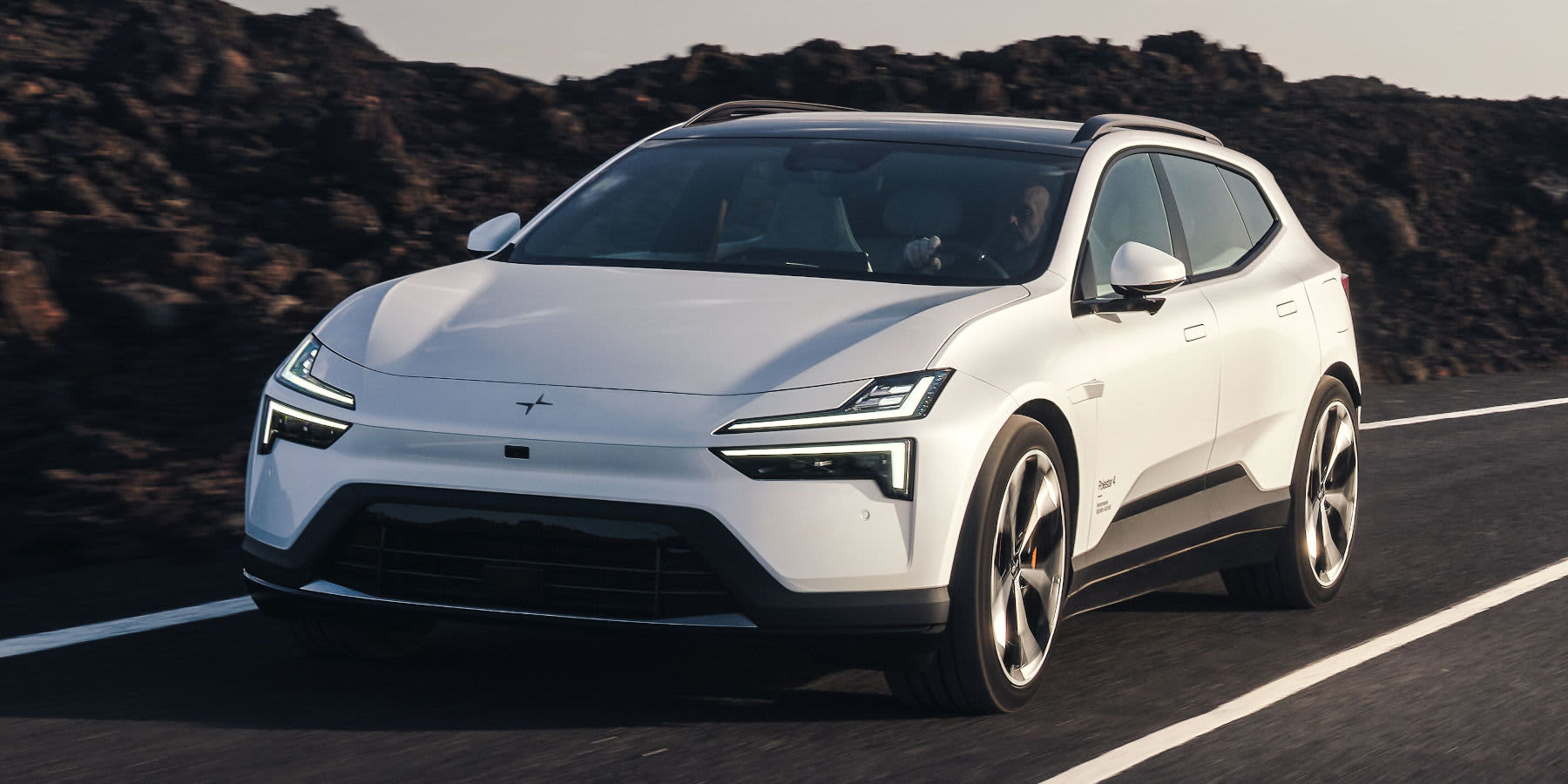 Ein weißer Polestar 3 Elektro-SUV fährt auf einer Straße vor einer dunklen Felslandschaft. Markant sind die geteilten LED-Scheinwerfer und das Panoramadach.