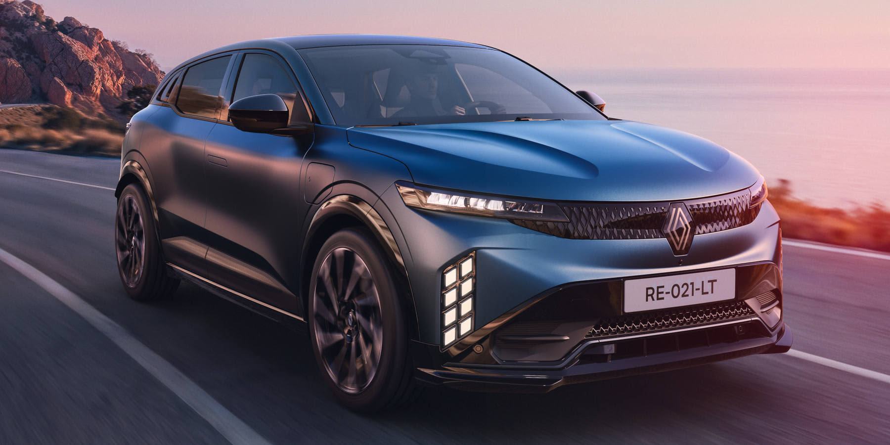 Ein blauer Renault Megane E-Tech elektrisch fährt bei Sonnenuntergang entlang einer malerischen Küstenstraße. Die markanten LED-Tagfahrlichter verleihen der dynamischen Front eine moderne Optik.