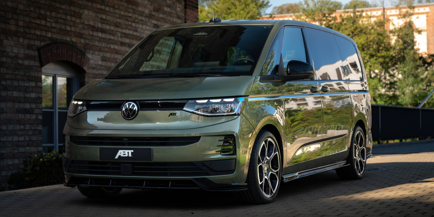 Ein olivgrüner VW Multivan von ABT steht vor einem Klinkergebäude. Prägend sind das ABT-Tuningpaket, Sportfelgen und der Frontsplitter.  