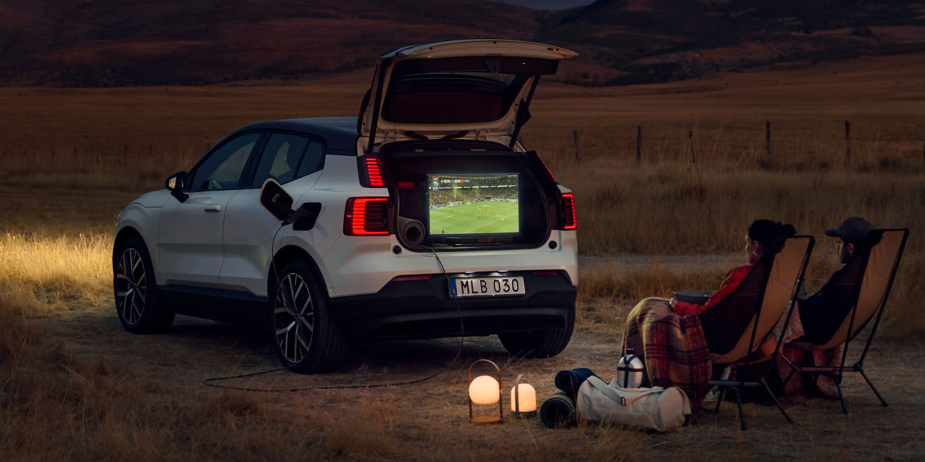 Weißer Volvo EX30 beim Camping, dessen Kofferraum einen Fernseher beim Fußballschauen mit Strom versorgt.
