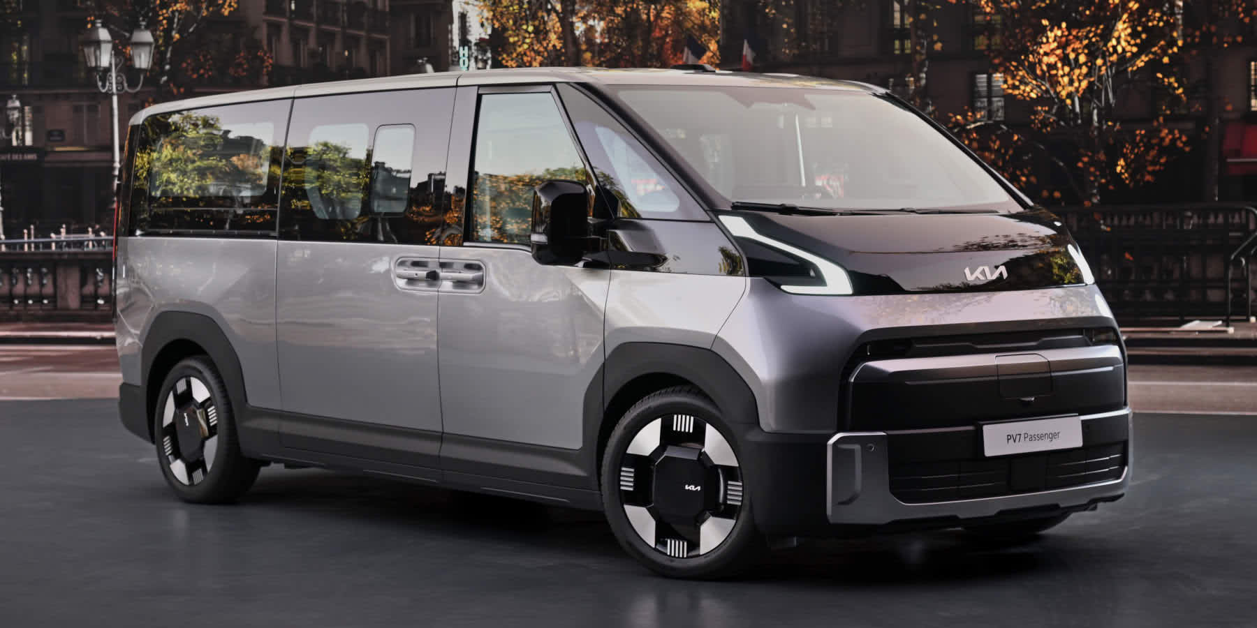 Ein silber-schwarzer Kia PV7 Passenger Van steht vor einer Allee. Prägend sind das futuristische Design und die markanten LED-Lichter.
