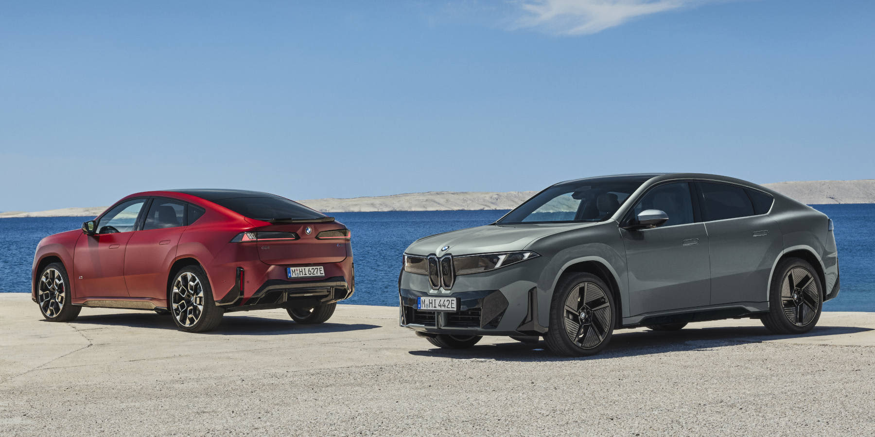 Zwei BMW iX4 stehen an einer Küstenstraße vor Meereskulisse. Prägend sind die schräge SUV-Coupé-Heckpartie, die schmale Niere und scharfe LED-Leuchten.   