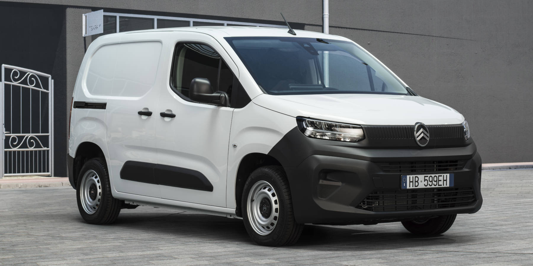 Ein weißer Citroën Berlingo Kastenwagen steht vor einem grauen Gebäude. Prägend sind der schwarze Kunststoff-Stoßfänger, kompakte Scheinwerfer und das neue Markenlogo.   